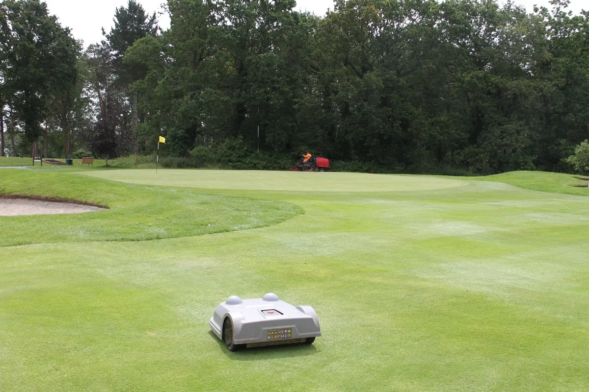 Robotic mower in the foreground with greenkeeping equipment in the distance