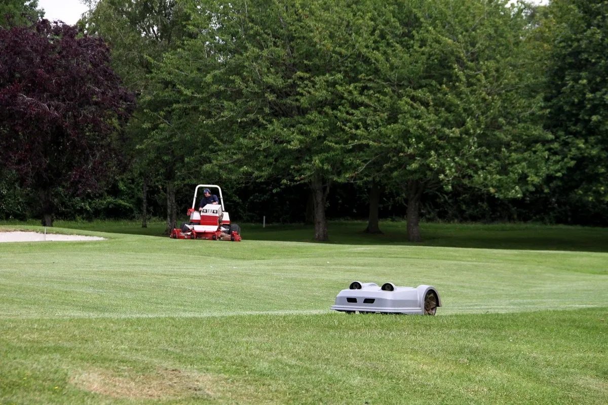 A robotic mower in the foreground with an operator on a conventional mower behind